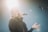 A person is singing passionately into a microphone while standing under bright stage lighting. The individual is wearing a dark sweater and has one hand extended outward. The lighting creates a dramatic effect with a noticeable glow around the head of the person.