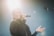 A person is singing passionately into a microphone while standing under bright stage lighting. The individual is wearing a dark sweater and has one hand extended outward. The lighting creates a dramatic effect with a noticeable glow around the head of the person.