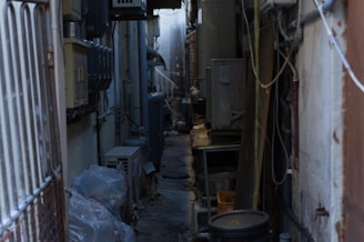 A narrow alleyway cluttered with various electrical and air conditioning units mounted on the walls. The passage is dimly lit and appears somewhat industrial. Plastic bags are piled on one side, and cables and pipes run along the walls.