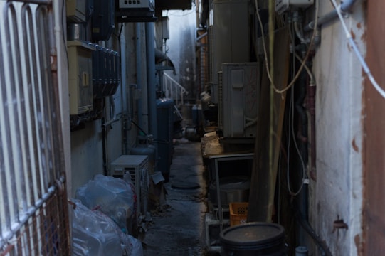 A narrow alleyway cluttered with various electrical and air conditioning units mounted on the walls. The passage is dimly lit and appears somewhat industrial. Plastic bags are piled on one side, and cables and pipes run along the walls.