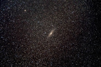 A dense field of countless stars scattered across a dark sky. At the center, there is a prominent bright galaxy with a faint halo. The stars range in brightness and appear as tiny white specks against the deep blackness of space.