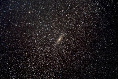 A dense field of countless stars scattered across a dark sky. At the center, there is a prominent bright galaxy with a faint halo. The stars range in brightness and appear as tiny white specks against the deep blackness of space.