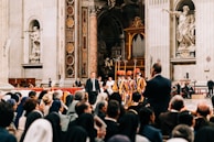 A large group of people is gathered inside an ornate church with intricate decorations and sculptures on the walls. Swiss Guards in traditional uniforms stand in formation, while seated audience members, some wearing religious garments, watch attentively.