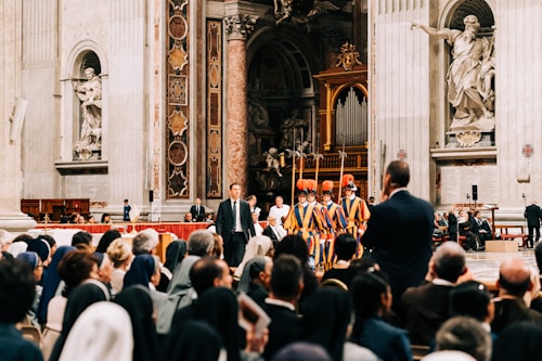 A large group of people is gathered inside an ornate church with intricate decorations and sculptures on the walls. Swiss Guards in traditional uniforms stand in formation, while seated audience members, some wearing religious garments, watch attentively.