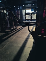 A dimly lit gym space with various fitness equipment including treadmills and weight machines. The sunlight creates long shadows on the floor, adding contrast to the scene.