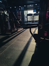 A dimly lit gym space with various fitness equipment including treadmills and weight machines. The sunlight creates long shadows on the floor, adding contrast to the scene.