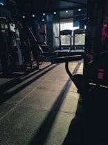 A dimly lit gym space with various fitness equipment including treadmills and weight machines. The sunlight creates long shadows on the floor, adding contrast to the scene.