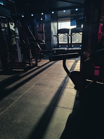 A dimly lit gym space with various fitness equipment including treadmills and weight machines. The sunlight creates long shadows on the floor, adding contrast to the scene.