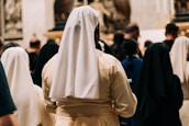 A group of nuns dressed in white and black habits is seen from behind, gathered among a crowd in what appears to be a religious or ceremonial setting with soft lighting and a blurred background.