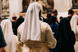 A group of nuns dressed in white and black habits is seen from behind, gathered among a crowd in what appears to be a religious or ceremonial setting with soft lighting and a blurred background.