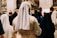 A group of nuns dressed in white and black habits is seen from behind, gathered among a crowd in what appears to be a religious or ceremonial setting with soft lighting and a blurred background.