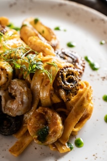 A close-up view of a gourmet pasta dish featuring fettuccine noodles. The dish includes a rich, creamy sauce with a golden-brown hue, complemented by pieces of shrimp and olives. Garnished with fresh herbs, the plate is artistically presented with small dots of green sauce around the edge.