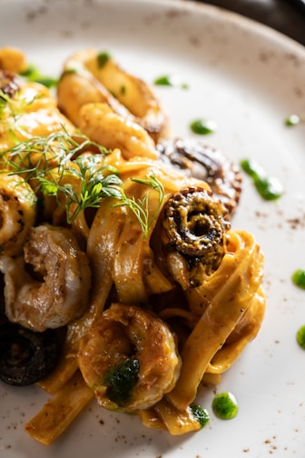 A close-up view of a gourmet pasta dish featuring fettuccine noodles. The dish includes a rich, creamy sauce with a golden-brown hue, complemented by pieces of shrimp and olives. Garnished with fresh herbs, the plate is artistically presented with small dots of green sauce around the edge.