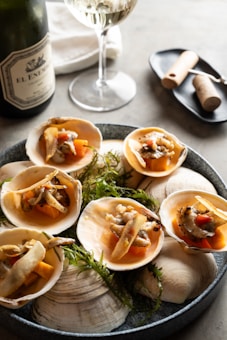 A beautifully arranged platter of seafood, featuring scallops served on half shells with garnishes of herbs and vegetables. Beside the plate is a glass of white wine and a wine bottle, all set on a table with a neutral, elegant background.