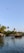 A tranquil waterway is surrounded by lush greenery and tall palm trees. Traditional houseboats float gently on the water. The sky is clear with a bird flying above.