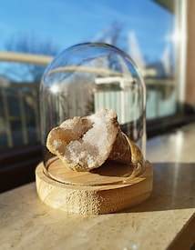 A rough, white crystal geode is displayed on a round wooden base. The geode is housed under a transparent glass dome. Sunlight casts soft shadows on the surface where the structure is placed, and a blurred view of a window and outdoor scenery is visible in the background.