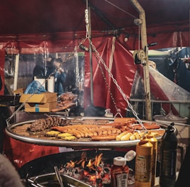 A large grilling setup with a variety of sausages and meats cooking over an open flame, surrounded by condiment bottles and a red tent backdrop. Chains hang from a metal frame, supporting the grill, and there are cardboard boxes and other supplies visible in the background.