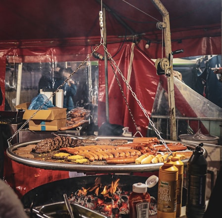 A large grilling setup with a variety of sausages and meats cooking over an open flame, surrounded by condiment bottles and a red tent backdrop. Chains hang from a metal frame, supporting the grill, and there are cardboard boxes and other supplies visible in the background.