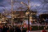 A festive outdoor scene with numerous people gathered around a fenced area decorated with Christmas lights. Bare trees are adorned with string lights, and there are small gingerbread-style houses. Large ornaments and candy-themed decorations are spread throughout the scene, creating a holiday atmosphere. It appears to be dusk, with a darkening sky providing contrast to the glowing lights.