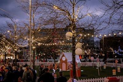 A festive outdoor scene with numerous people gathered around a fenced area decorated with Christmas lights. Bare trees are adorned with string lights, and there are small gingerbread-style houses. Large ornaments and candy-themed decorations are spread throughout the scene, creating a holiday atmosphere. It appears to be dusk, with a darkening sky providing contrast to the glowing lights.