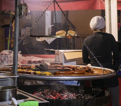 A street food stall with a large round grill suspended above hot coals, covered with a variety of sausages and meats. A person wearing a gray beanie stands next to the grill, overseeing the cooking. Buns are placed on a shelf above the grill, along with a cardboard box. In the background, there are bottled drinks and other supplies.