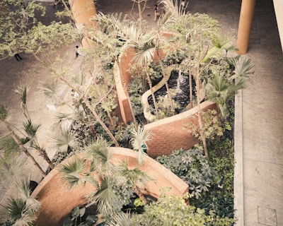 An aerial view of a landscaped area featuring a series of curved brick walls enclosing lush green vegetation. Tall trees with narrow trunks and various types of leafy plants fill the space. The design incorporates a small pond or water feature that adds a tranquil element. The surrounding area includes tiled flooring, and a few people are seen walking along the paths.