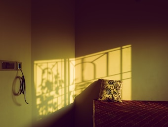 A room corner featuring sunlight streaming through a window, casting intricate shadows on the wall. A power outlet is visible with a black cable hanging beside it. A bed is partially visible with a patterned pillow leaning against the wall.