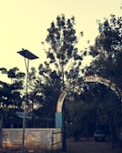 An arched entrance with text in a regional language leads to a tree-filled area. A metal gate and concrete wall with painted instructions are visible. A lamppost with a solar panel stands nearby, and a car is parked in the background.