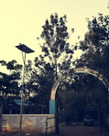 An arched entrance with text in a regional language leads to a tree-filled area. A metal gate and concrete wall with painted instructions are visible. A lamppost with a solar panel stands nearby, and a car is parked in the background.