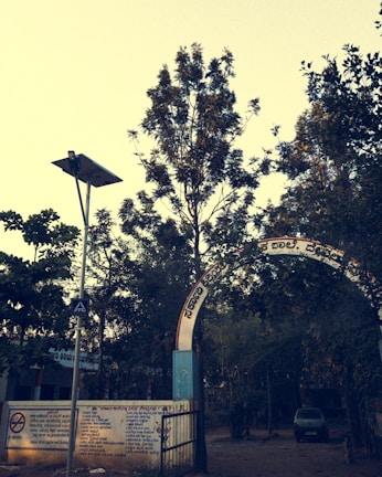 An arched entrance with text in a regional language leads to a tree-filled area. A metal gate and concrete wall with painted instructions are visible. A lamppost with a solar panel stands nearby, and a car is parked in the background.