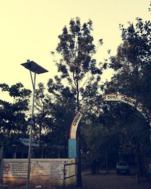 An arched entrance with text in a regional language leads to a tree-filled area. A metal gate and concrete wall with painted instructions are visible. A lamppost with a solar panel stands nearby, and a car is parked in the background.