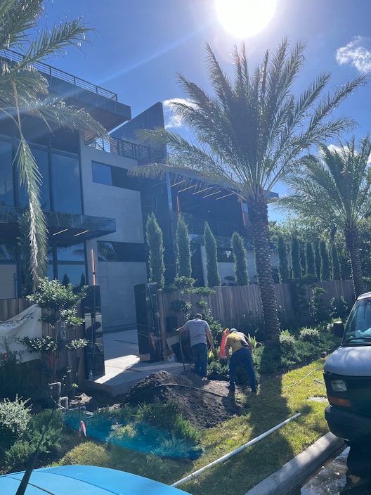 Two workers are engaged in landscaping activities in front of a modern building. The scene is sunny and features several palm trees and well-maintained greenery. A pile of soil is present, and a white van is parked nearby.