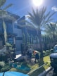 Two workers are engaged in landscaping activities in front of a modern building. The scene is sunny and features several palm trees and well-maintained greenery. A pile of soil is present, and a white van is parked nearby.