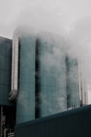 A modern industrial building with cylindrical structures and large metal pipes surrounded by steam or fog. The architecture features a teal-colored surface, lending a sleek and modern appearance, while the atmosphere is somewhat mysterious due to the misty surroundings.