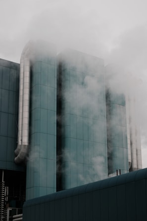 A modern industrial building with cylindrical structures and large metal pipes surrounded by steam or fog. The architecture features a teal-colored surface, lending a sleek and modern appearance, while the atmosphere is somewhat mysterious due to the misty surroundings.