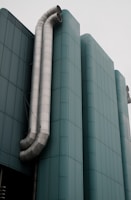 A modern building with sleek, curved lines and a teal facade. Two large metallic pipes run vertically along the side, contrasting against the smooth architectural design.