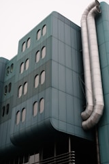 A modern building facade featuring teal-colored panels and multiple oval windows. Large metallic ventilation pipes curve down the side, adding an industrial element to the sleek architecture.