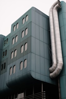 A modern building facade featuring teal-colored panels and multiple oval windows. Large metallic ventilation pipes curve down the side, adding an industrial element to the sleek architecture.