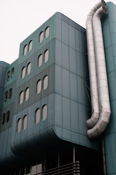 A modern building facade featuring teal-colored panels and multiple oval windows. Large metallic ventilation pipes curve down the side, adding an industrial element to the sleek architecture.
