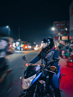 Motorcycle rider adjusting a compact intercom device on their helmet with city lights behind.