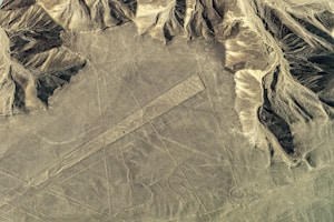 An aerial view of a desert landscape with large, mysterious geometric lines and shapes etched into the ground. These are set against a backdrop of rugged, eroded hills and rock formations.
