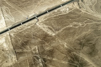 An aerial view of a desert landscape featuring large, ancient geoglyphs, including a figure that resembles a person with long limbs. A road cuts through the scene, indicating a mix of natural and human-made elements in the environment.