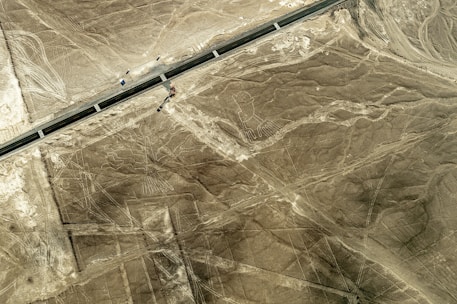 An aerial view of a desert landscape featuring large, ancient geoglyphs, including a figure that resembles a person with long limbs. A road cuts through the scene, indicating a mix of natural and human-made elements in the environment.