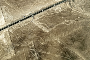 An aerial view of a desert landscape featuring large, ancient geoglyphs, including a figure that resembles a person with long limbs. A road cuts through the scene, indicating a mix of natural and human-made elements in the environment.