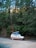 A camper van with a raised roof parked on a dirt path, surrounded by dense, tall evergreen trees. The setting is serene and natural, suggesting a location in a forest or wooded area.