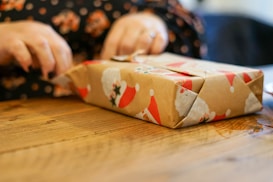 Hands are wrapping a gift on a wooden table using festive wrapping paper with images of Santa Claus. The focus is on the gift being wrapped, with a blurred background.