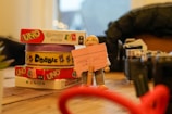 A wooden table displaying a stack of card games including 'Uno' and 'Dobble', with a small figurine holding a pink note with handwritten text. To the side, various objects including parts of a camera and blurred items in the foreground and background.