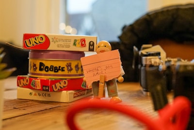 A wooden table displaying a stack of card games including 'Uno' and 'Dobble', with a small figurine holding a pink note with handwritten text. To the side, various objects including parts of a camera and blurred items in the foreground and background.