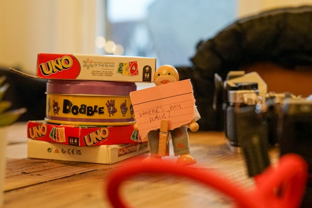 A wooden table displaying a stack of card games including 'Uno' and 'Dobble', with a small figurine holding a pink note with handwritten text. To the side, various objects including parts of a camera and blurred items in the foreground and background.