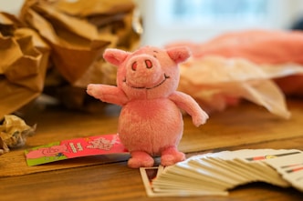 A small, plush pink pig toy is standing on a wooden surface. To one side, near the toy, there is a colorful label with illustrations. In the foreground, a spread of playing cards is visible, partially fanned out. The background features crumpled brown paper and a blurry, soft pink object that seems to be fabric or tissue.